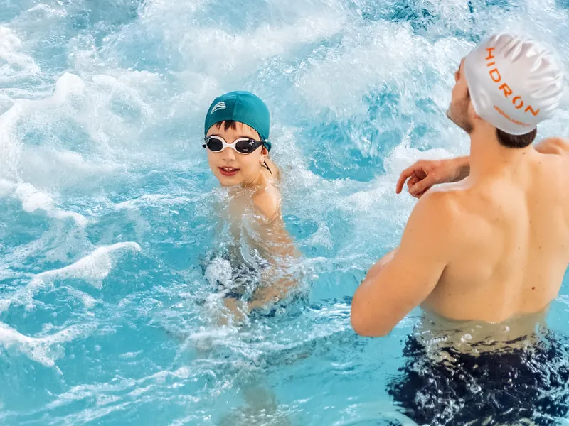 Un bambino con gli occhialini ride in acqua, con un adulto accanto