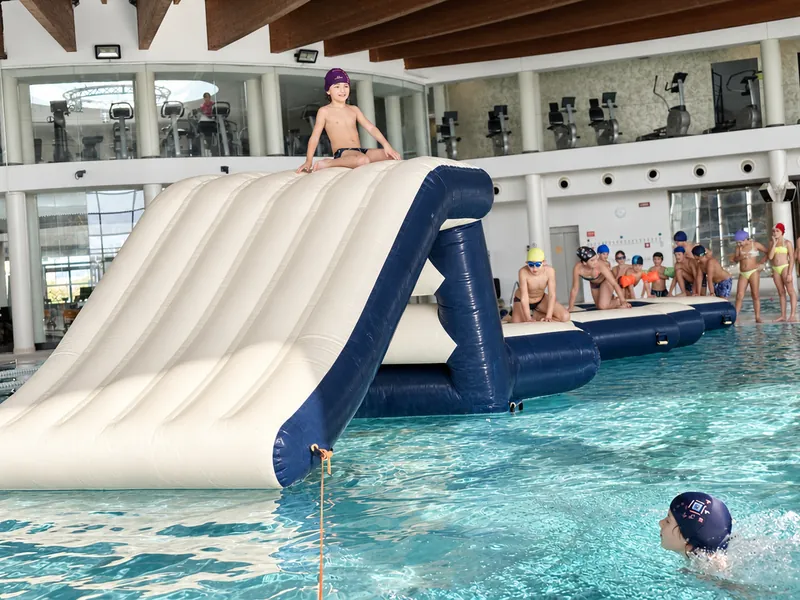 Un bambino seduto in cima a uno scivolo gonfiabile bianco e blu, in acqua, e altri bambini che aspettano il turno sulle piattaforme
